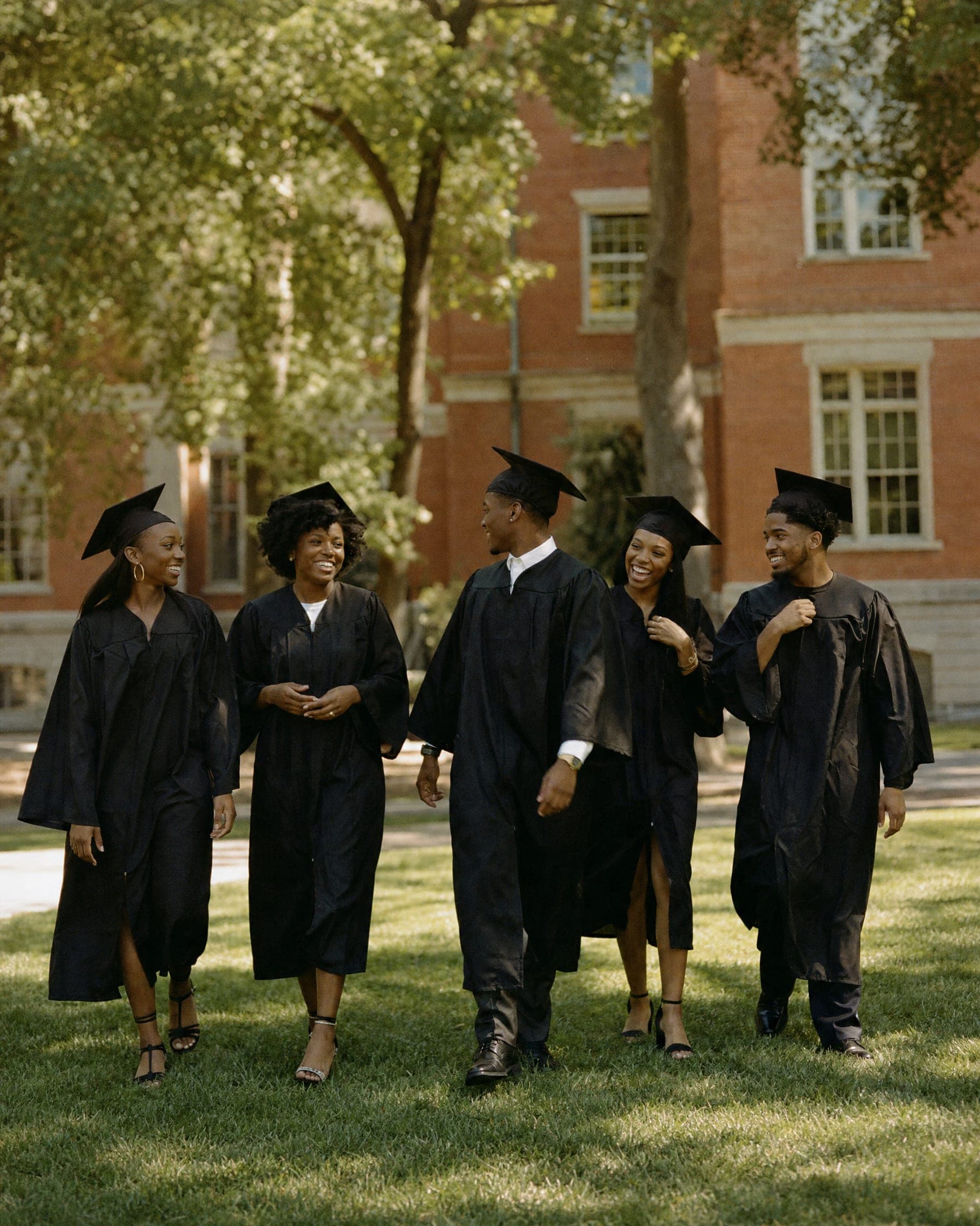 Five graduates in caps and gowns walk together across a sunny campus lawn, laughing.