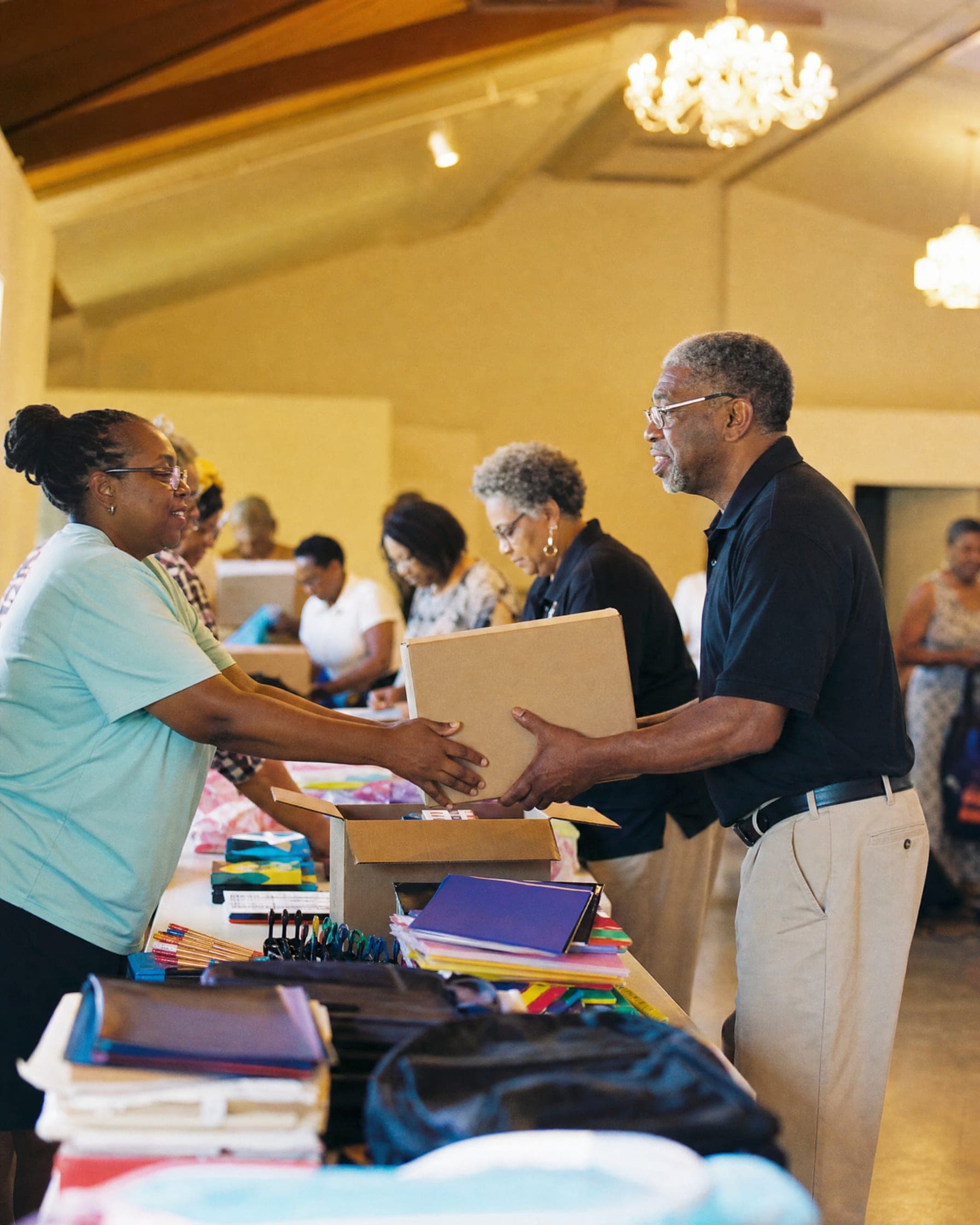 Volunteers of different ages set up tables of donated school supplies in a fellowship hall.