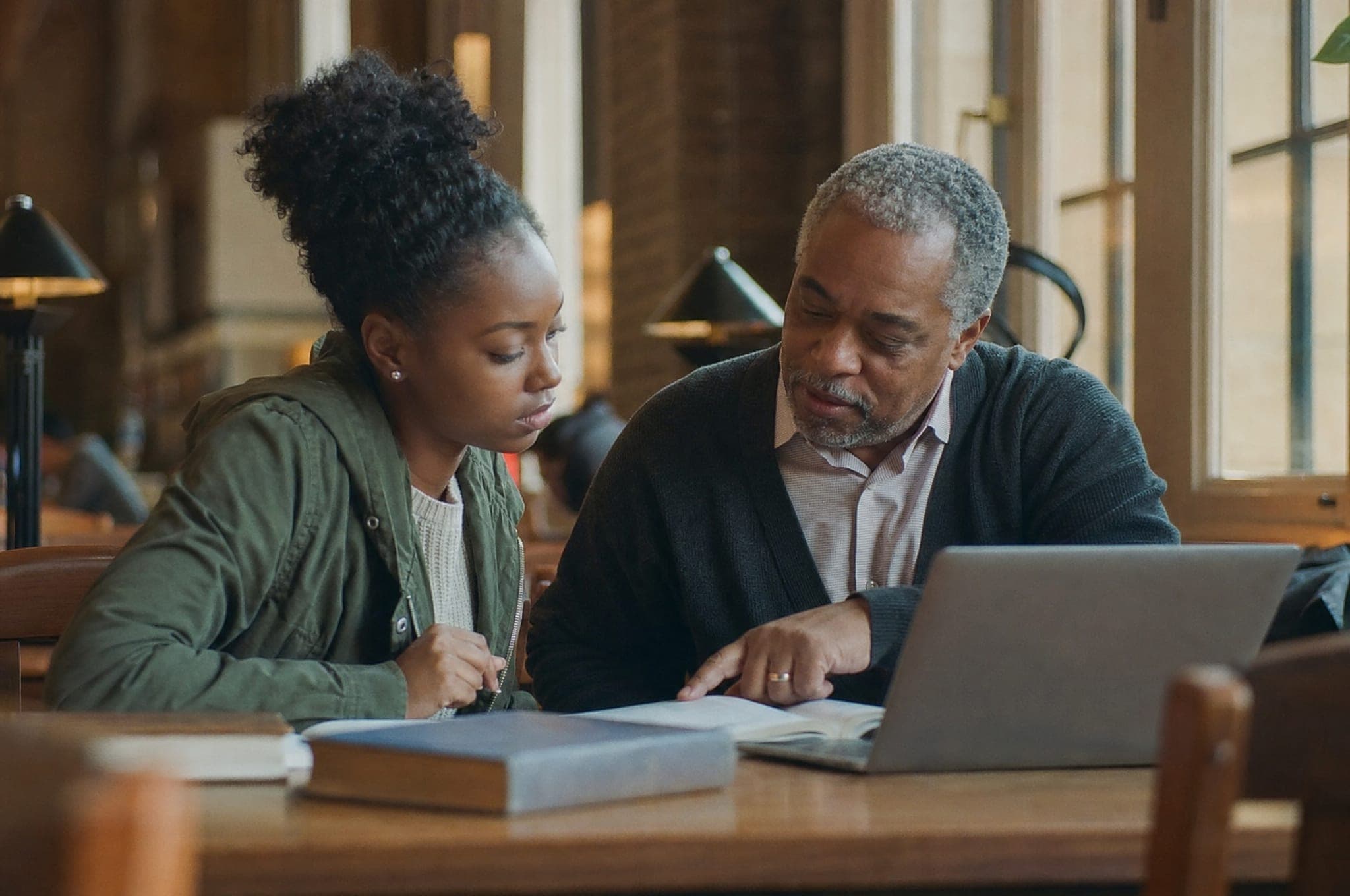 A college student studies at a library table while an older mentor beside her points to a passage in her textbook.