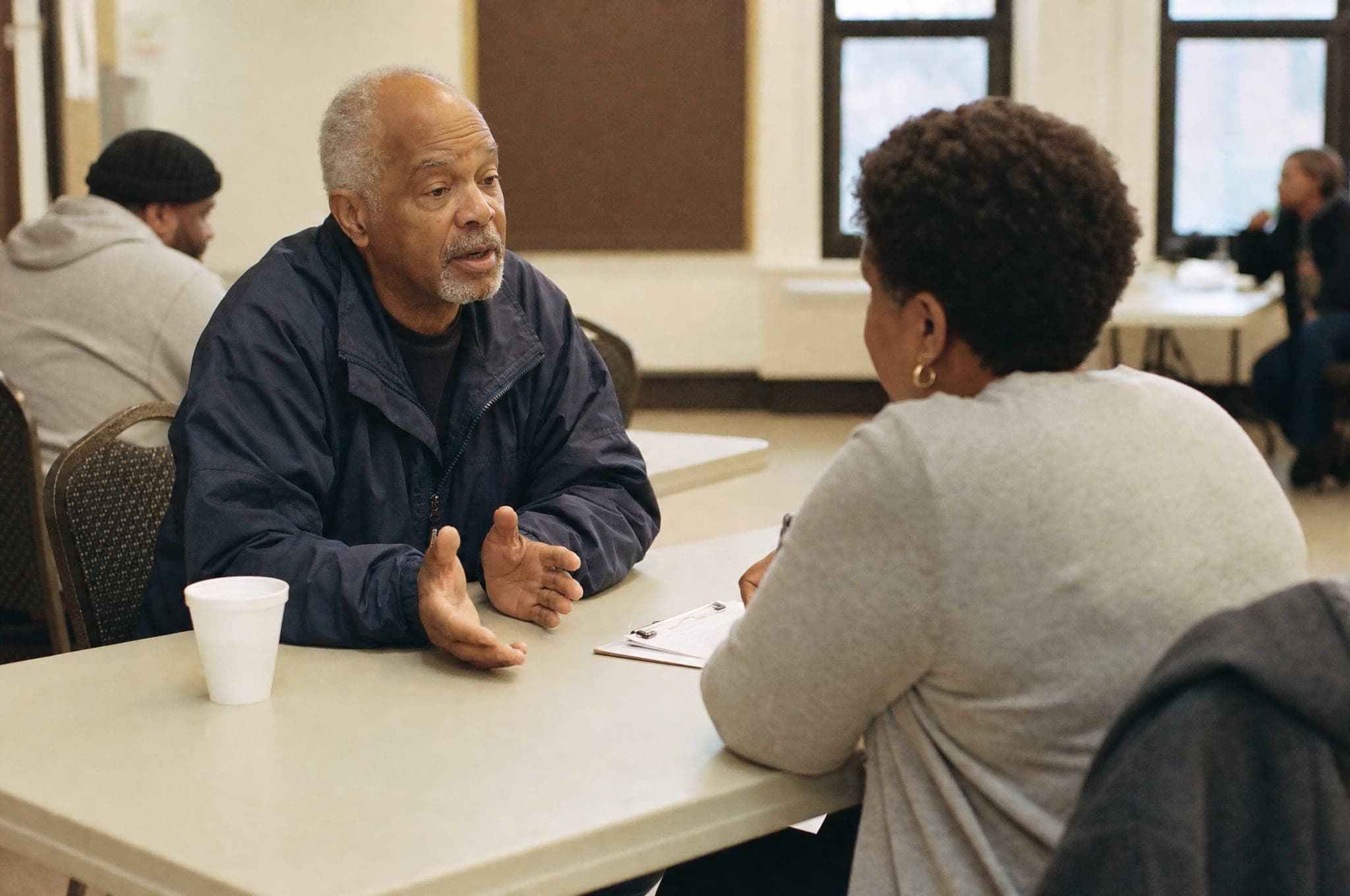 A veteran in his sixties talks across a table in a community center while a volunteer, seen from behind, listens with a clipboard on the table between them.